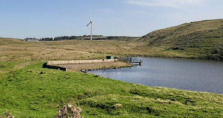 A body of water surrounded by green grass fields and a small hill on the right hand side. A wind turbine is in the distance and blue sky is overhead.