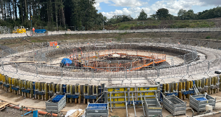 Construction site with a circular foundation structure surrounded by scaffolding and construction materials, under a partly cloudy sky.