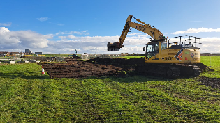 An excavator is digging into a grassy field under a partly cloudy sky. The machine is removing soil to create a trench, with open green space and distant buildings visible in the background.