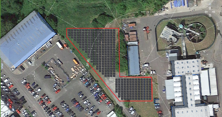 An aerial view of the location of solar panels. A white office building is on the right and another blue building is on the left with the solar panels in the middle. Trees and greenery are at the top with a car park also on the left.