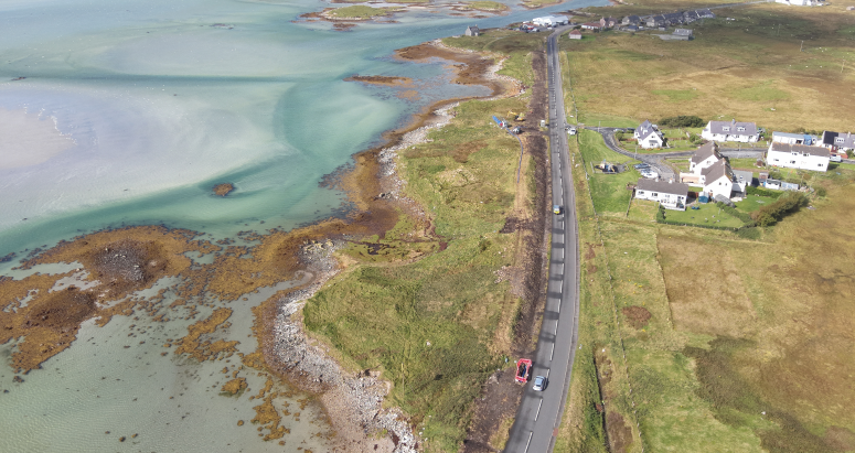 Aerial view of a coastal road in Benbecula, Scotland, with adjacent houses and clear turquoise waters alongside rocky shorelines.