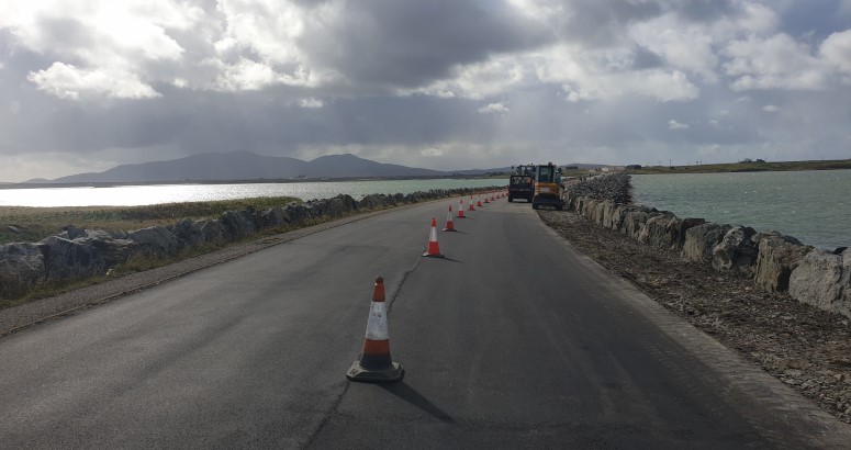 Road construction in progress along a coastal road with traffic cones lining one side and machinery ahead under a cloudy sky.