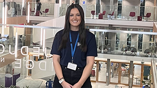  Trainee Manager Holly Henderson in front of a glass awards cabinet promoting Apprenticeship Week