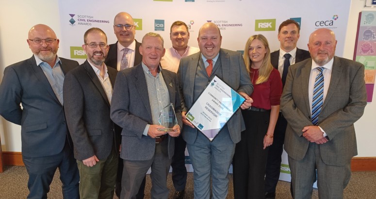Group of nine people posing with an award at an event. Some are smiling and holding a certificate and a trophy. The setting suggests a professional or corporate event.