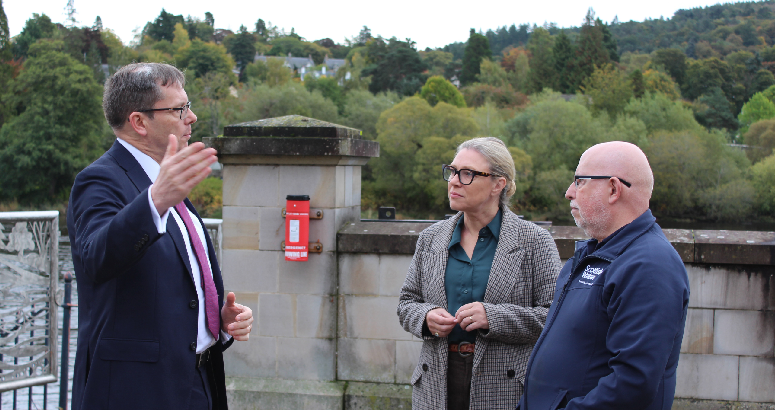 A man stands to the left of the picture, facing into the frame, talking to a female and a male who are looking at home. Behind them is a shoulder-height concrete wall, and trees in the background.