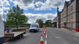 A row of traffic cones is placed down the centre of a two-way road. On the right hand side of the picture there is a row of buildings. On the left hand side, there are vehicles parked in the coned-off area.