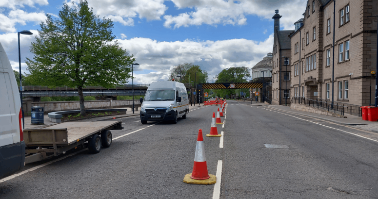 A row of traffic cones is placed down the centre of a two-way road. On the right hand side of the picture there is a row of buildings. On the left hand side, there are vehicles parked in the coned-off area.