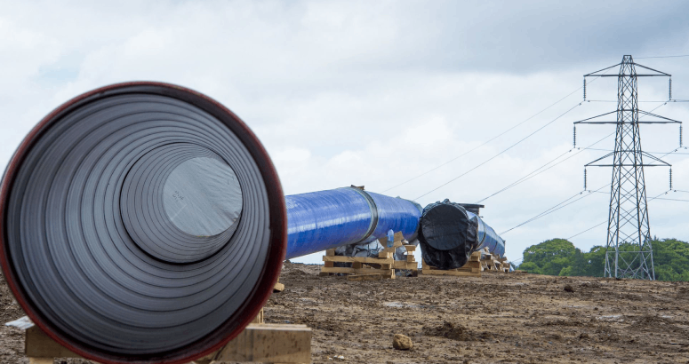 A ground-level photograph of an open water pipe upon the ground. The pipe is blue. In the background is an electricity pylon.