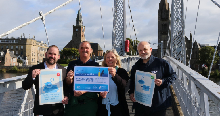 A group of four people stand outdoors on a bridge. They are holding blue and white posters. There are building in the background and a cloudy sky.