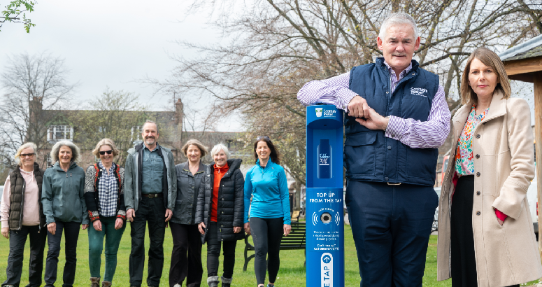 A group of people stand in the background on a grassed area, while in the foreground two people stand beside a blue Scottish Water top up tap, with one of them resting their arm and elbow on it.