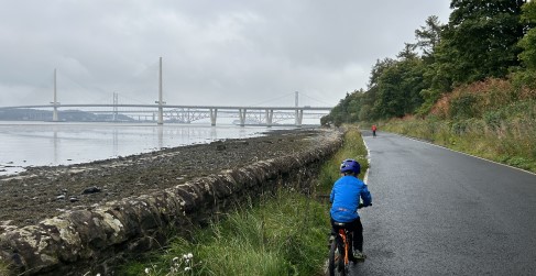 Two children in bright clothing cycling on tarmac road with bridges in the background