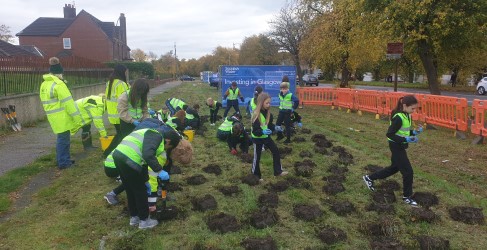 Group of volunteers wearing safety vests planting trees by the roadside, with a sign reading "Investing in Glasgow".