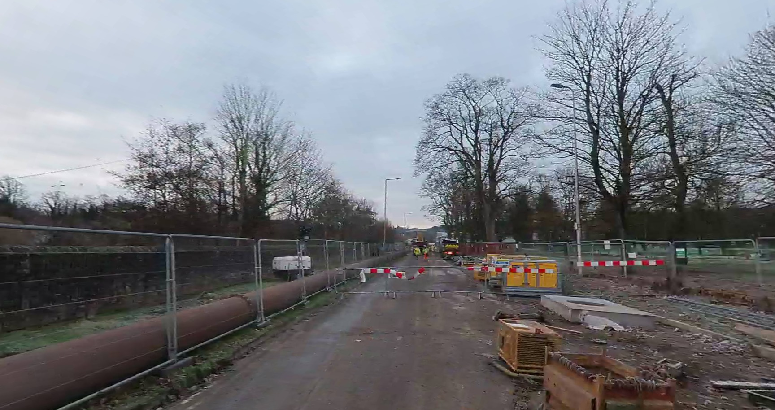 Construction site on a partially closed Shore Road with barriers and fencing; pipes and construction materials are visible along the road, with trees and overcast skies in the background.