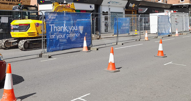 Busy street view showing a fenced off sewer repair works site with Thank You for Your Patience signage